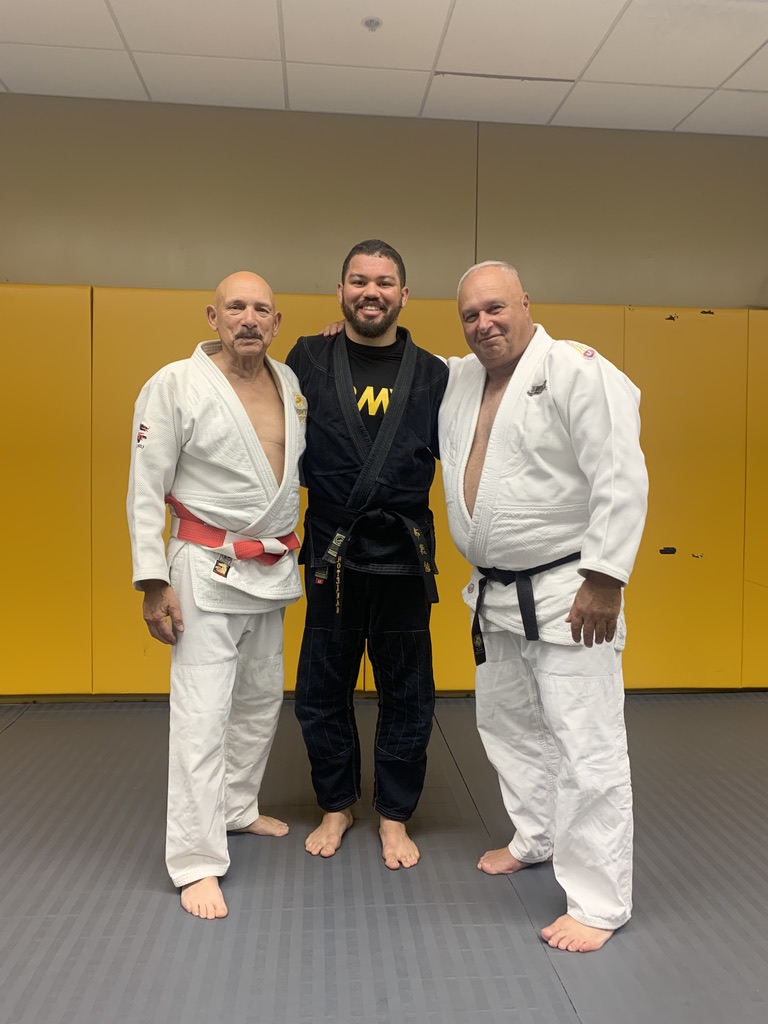 Christian Pierce in a black gi standing with his two Judo teachers on the mats